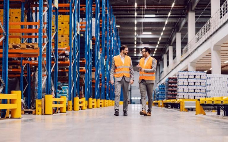 Two workers discussing in warehouse aisle.